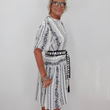 A woman with short blonde hair and glasses wearing a white short-sleeve dress with black vertical stripes and black floral embroidery, standing against a plain white wall.
