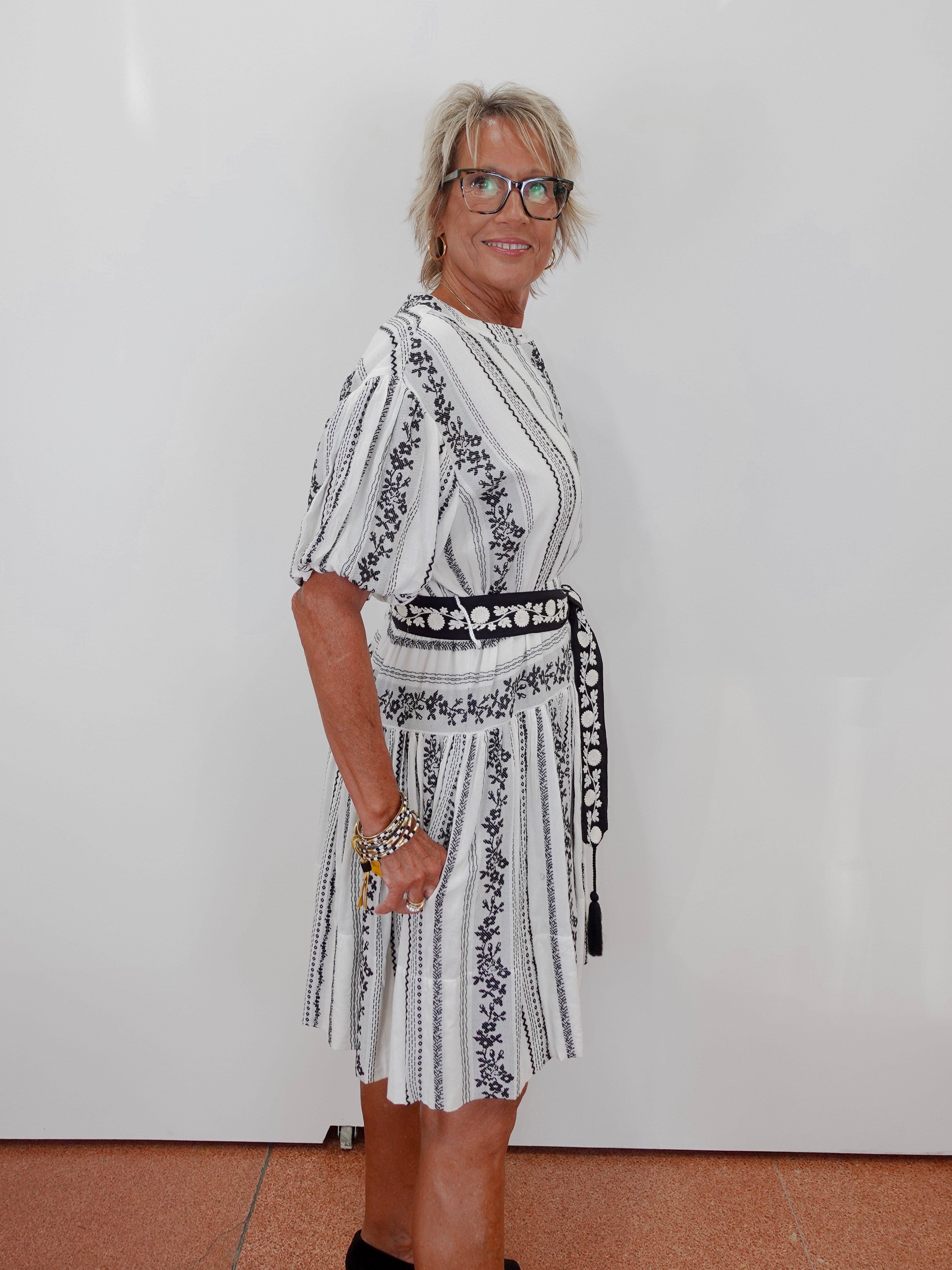 A woman with short blonde hair and glasses wearing a white short-sleeve dress with black vertical stripes and black floral embroidery, standing against a plain white wall.