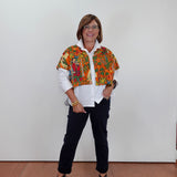 A woman wearing a colorful patterned cropped jacket over a white button-down shirt, paired with dark pants and gray wedge sandals, standing against a plain white wall.
