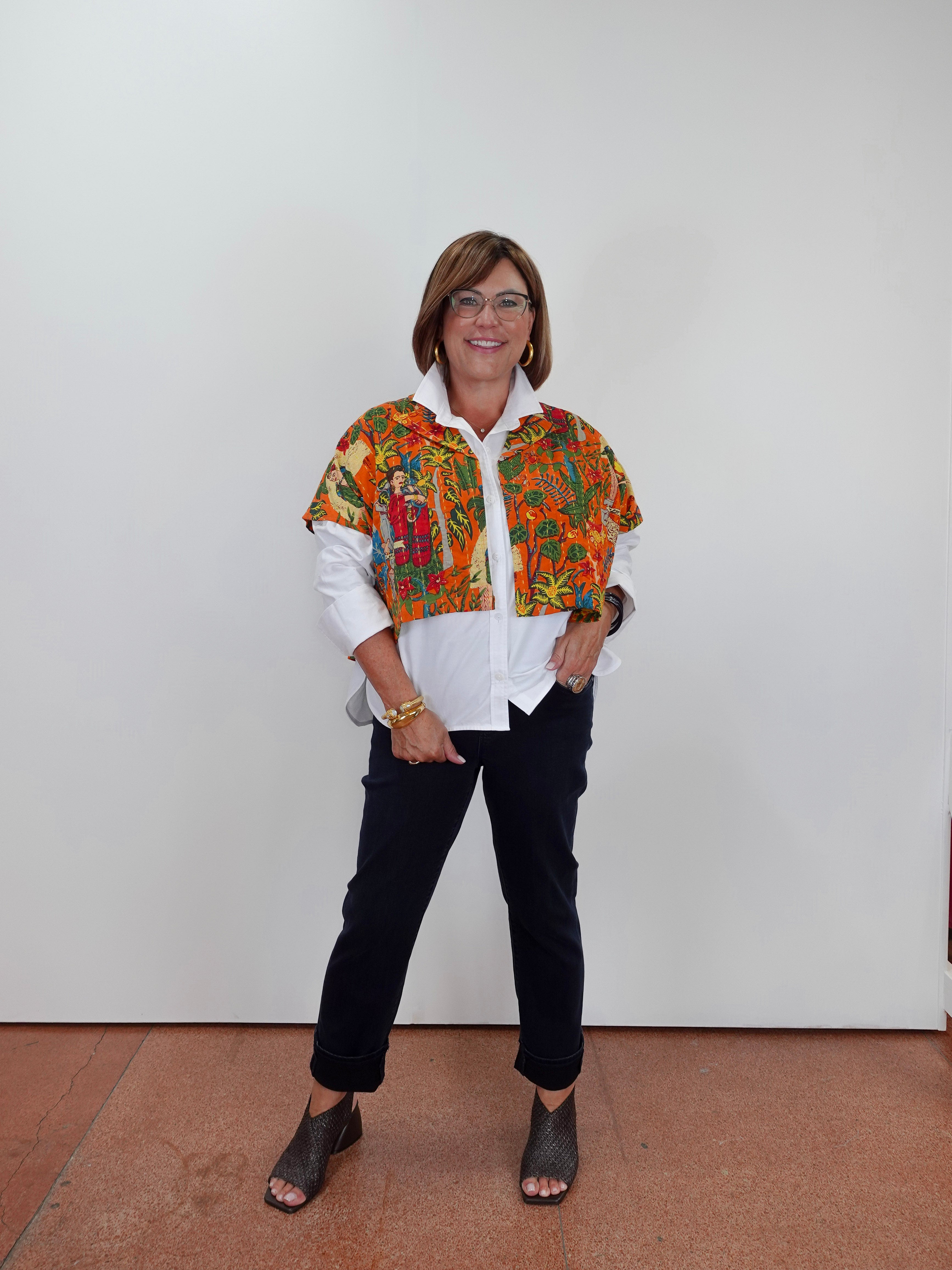 A woman wearing a colorful patterned cropped jacket over a white button-down shirt, paired with dark pants and gray wedge sandals, standing against a plain white wall.