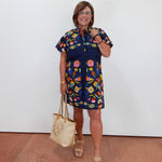 A woman modeling a short-sleeve navy blue dress with a colorful floral and butterfly print, holding a beige tote bag and wearing beige sandals against a plain white wall.