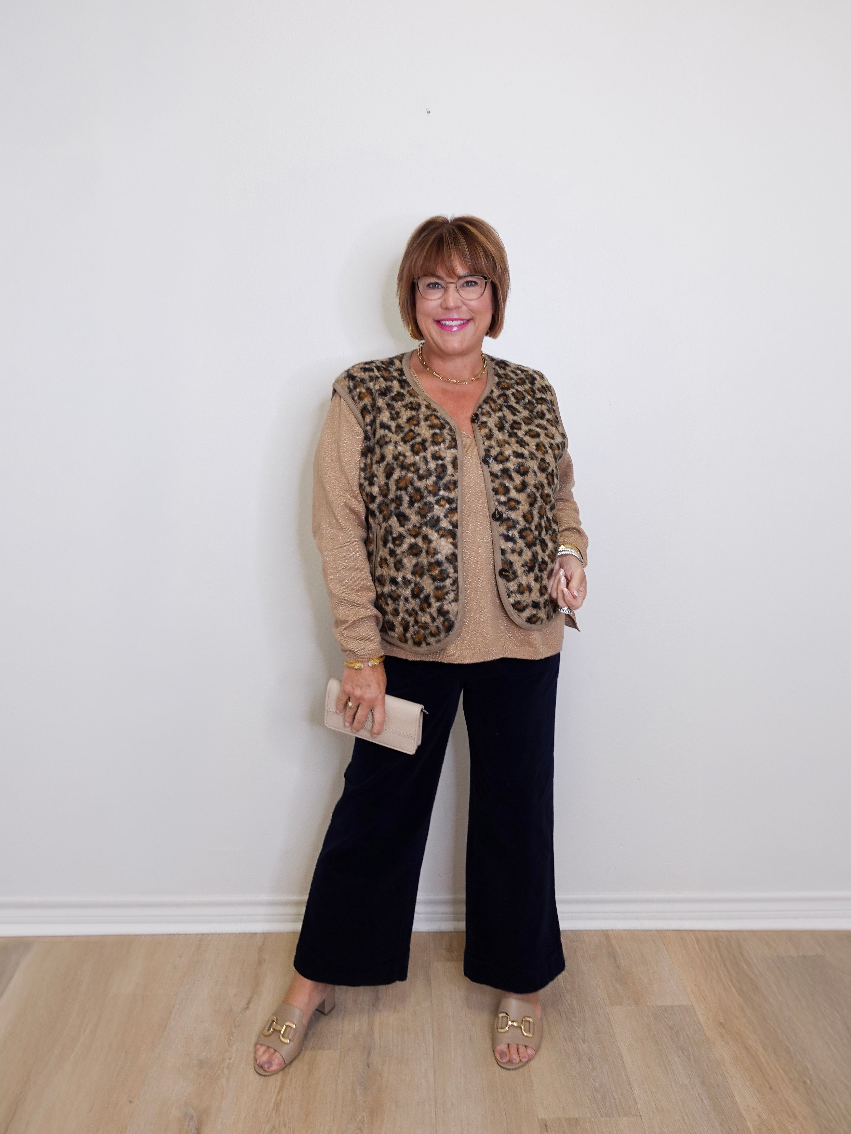 Woman wearing a leopard print vest over a beige sweater with black pants, standing against a white wall.