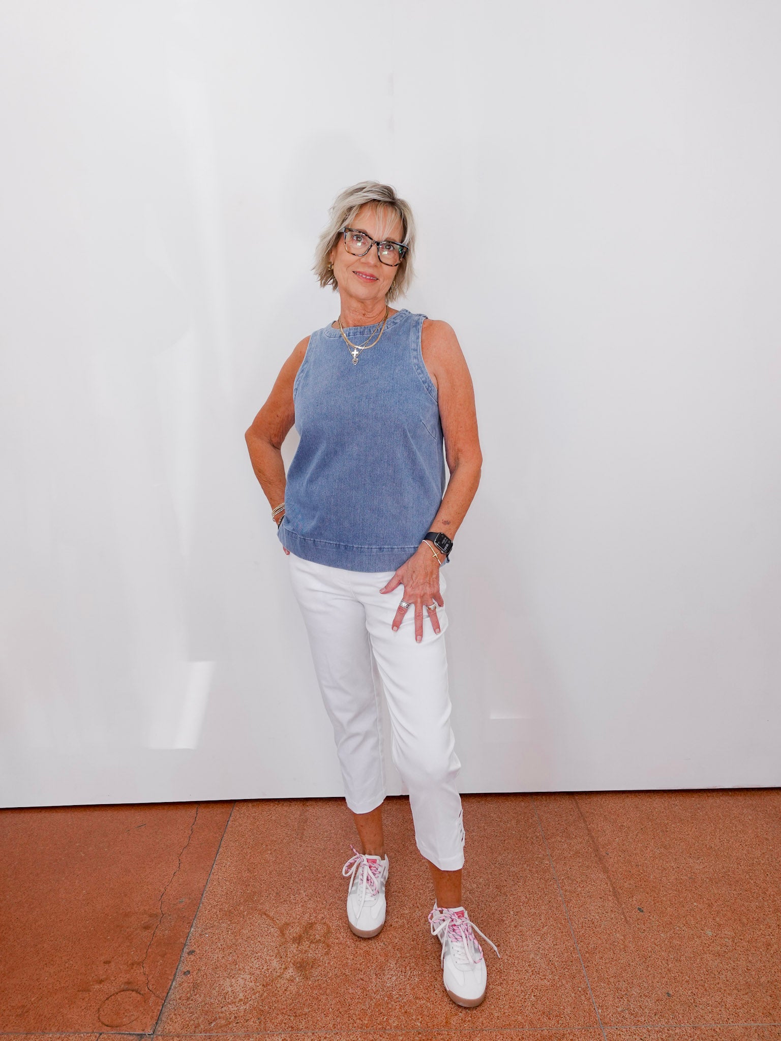 Woman wearing a blue sleeveless top and white pants against a white wall.