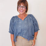 Woman wearing a blue blouse with floral patterns and beige pants against a plain background