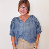 Woman wearing a blue blouse with floral patterns and beige pants against a plain background