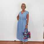 Woman in a blue dress holding a colorful bag against a white wall.