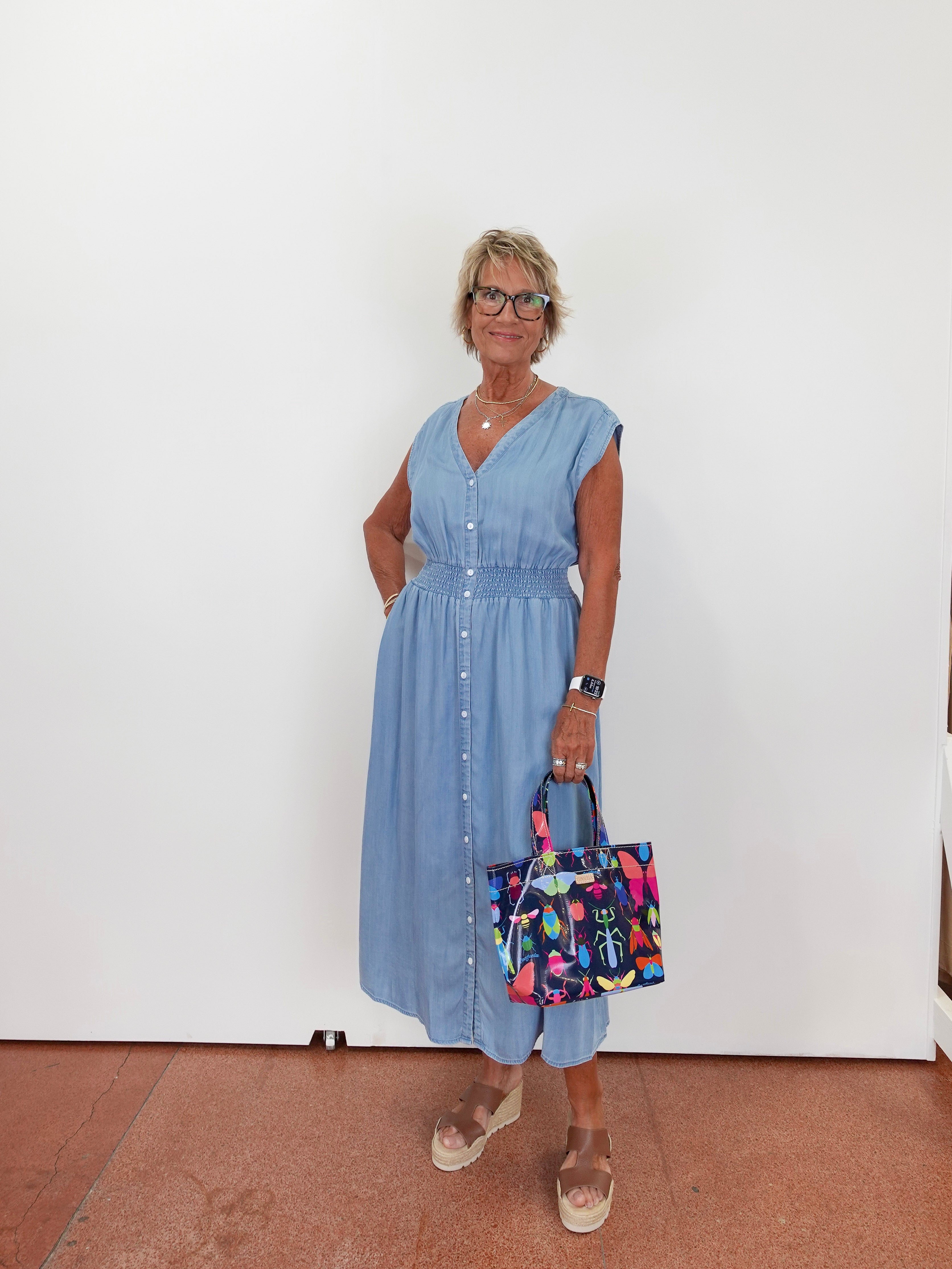 Woman in a blue dress holding a colorful bag against a white wall.