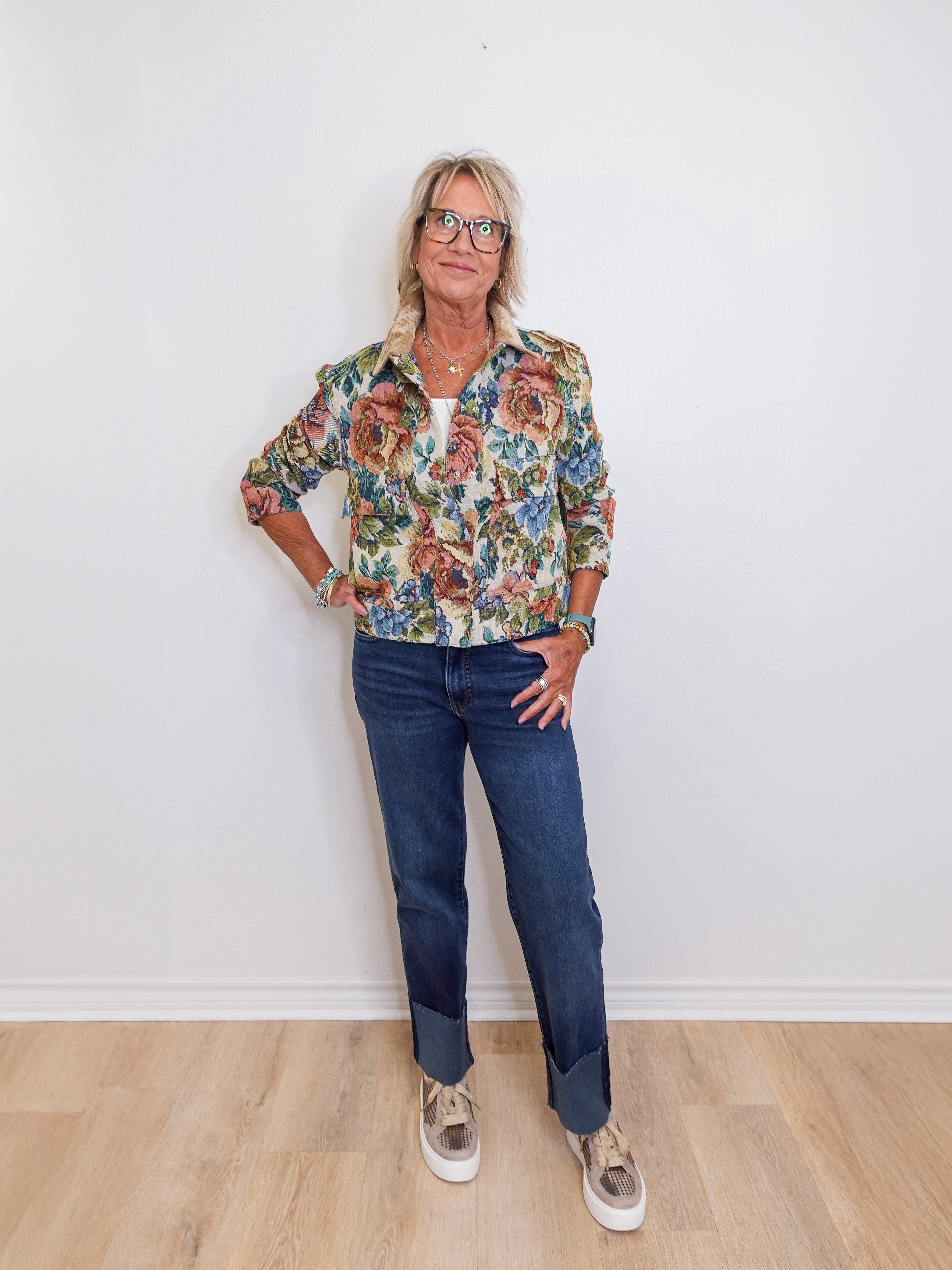 Woman wearing a floral shirt and jeans standing against a white wall.