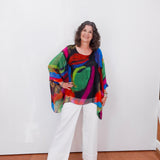 Woman wearing a colorful, patterned top and white pants against a white wall.