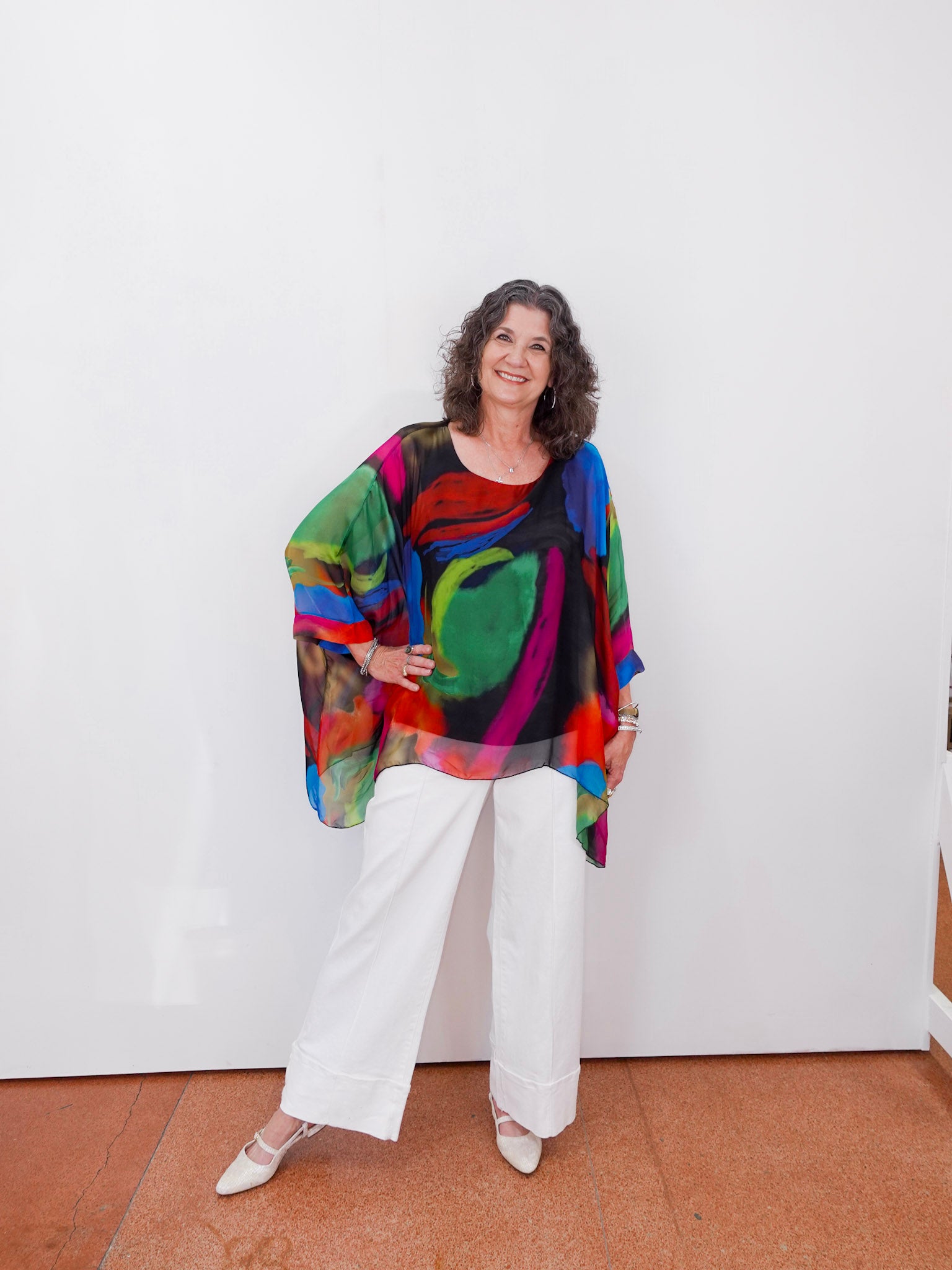 Woman wearing a colorful, patterned top and white pants against a white wall.