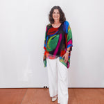 Woman wearing a colorful top and white pants standing against a white wall.