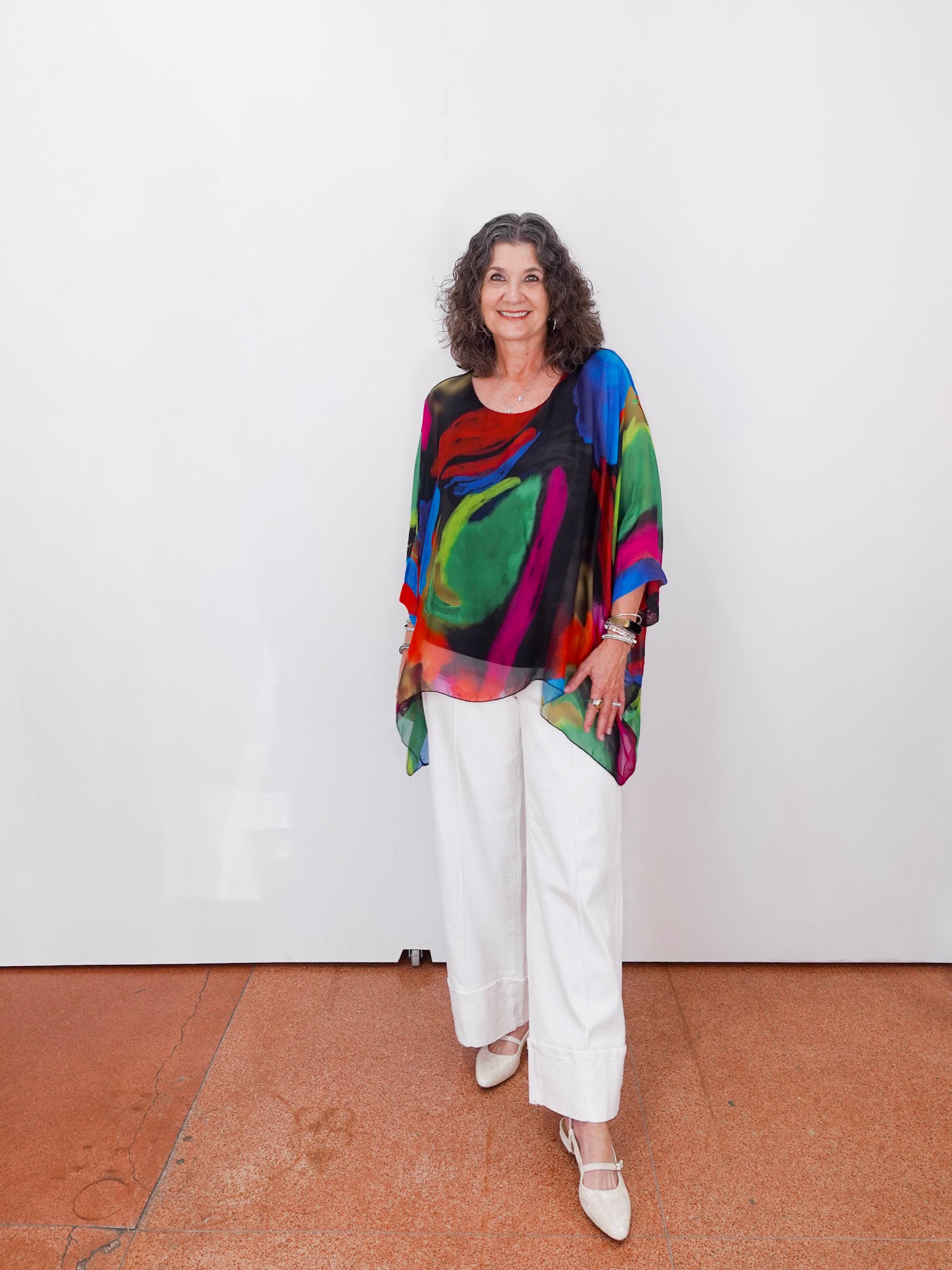 Woman wearing a colorful top and white pants standing against a white wall.