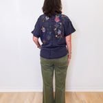 Person wearing a dark blue shirt with floral embroidery and green pants against a white wall.
