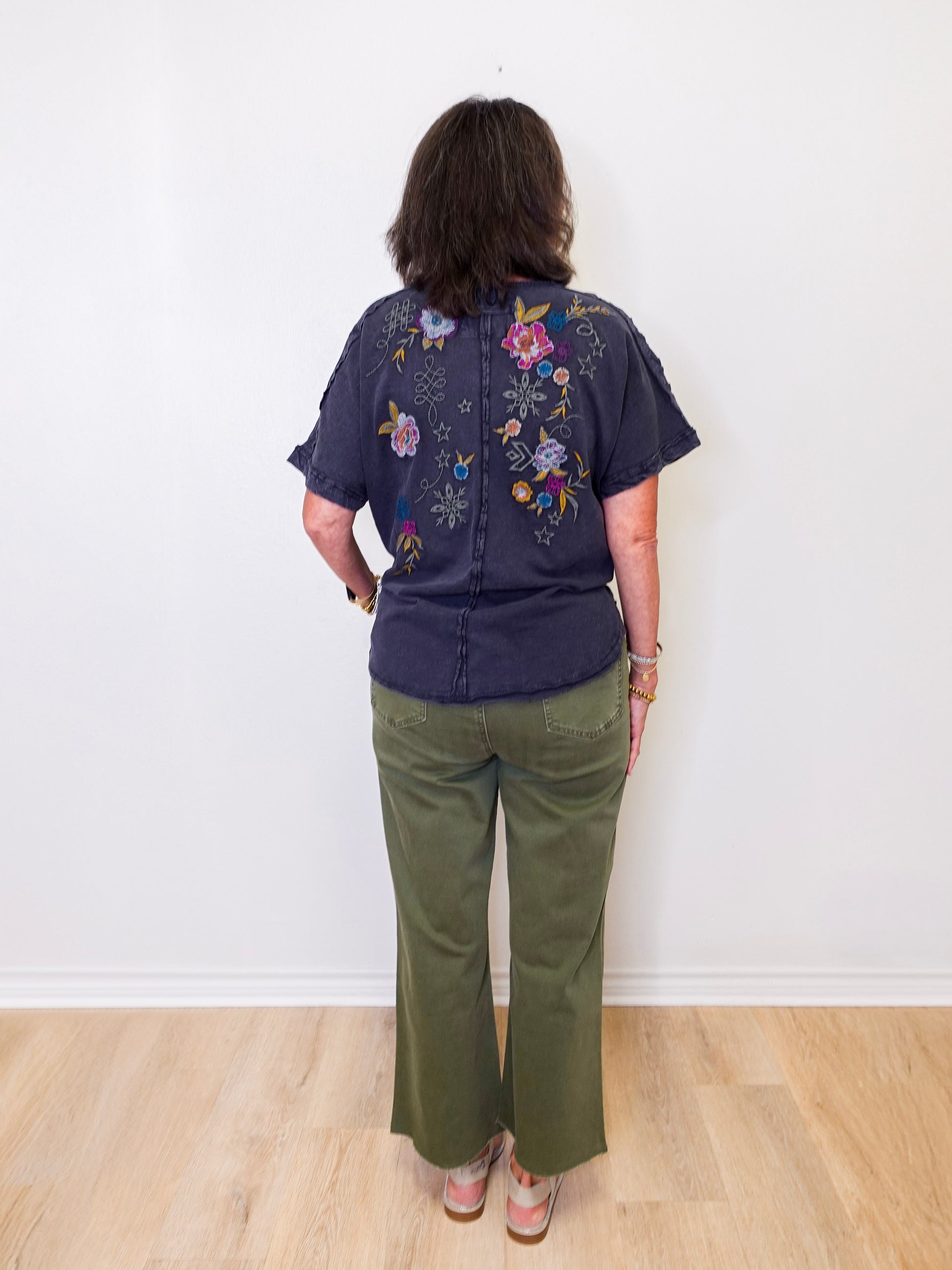 Person wearing a dark blue shirt with floral embroidery and green pants against a white wall.