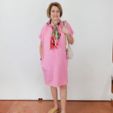 Woman wearing a pink dress with a colorful scarf and sandals, standing against a white wall.
