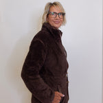 Person wearing a brown jacket and glasses against a white wall