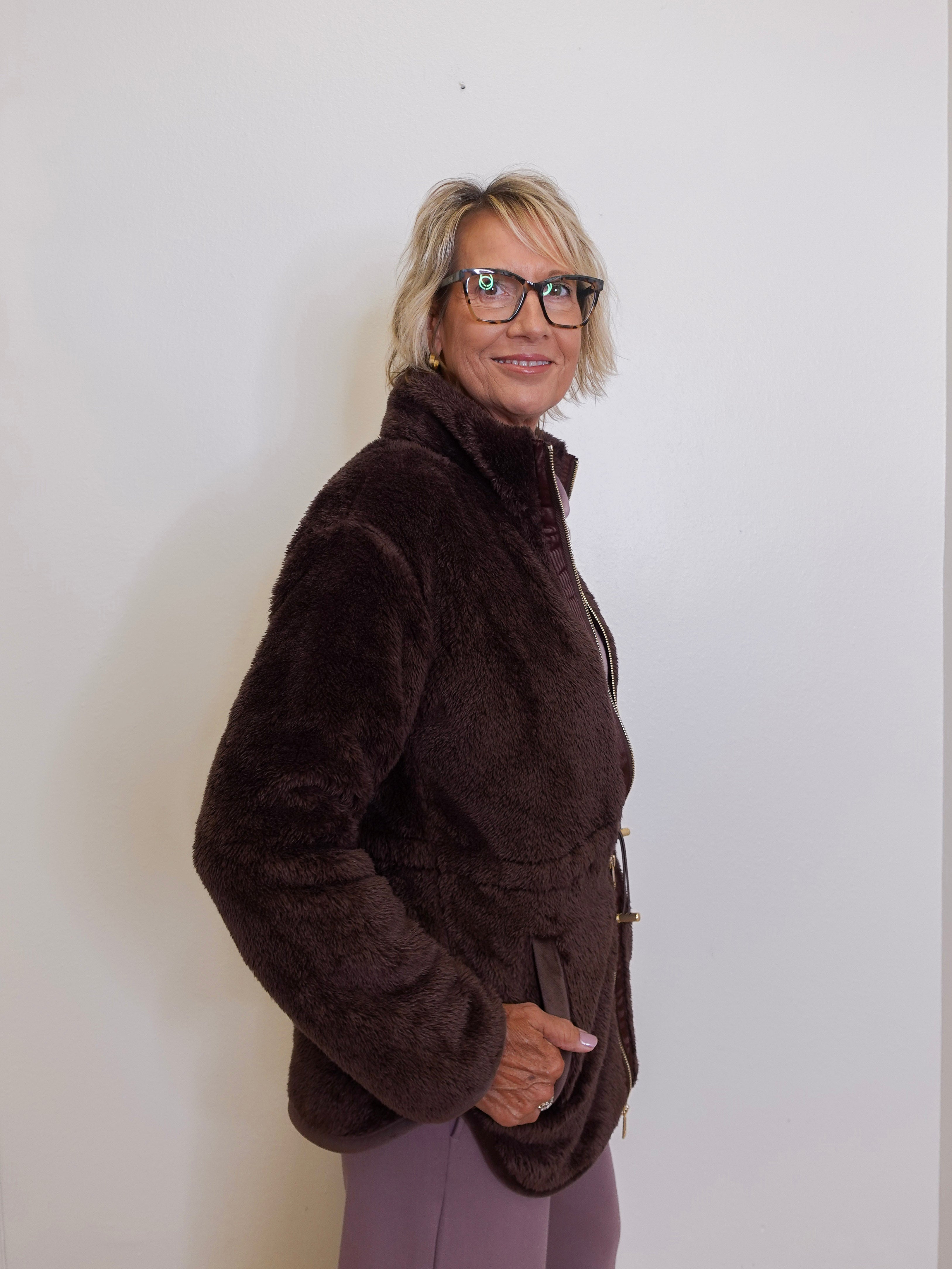 Person wearing a brown jacket and glasses against a white wall