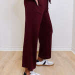 Person wearing burgundy pants and white sneakers with black stripes on a wooden floor.