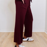 Person wearing burgundy pants and white sneakers with black stripes on a wooden floor.