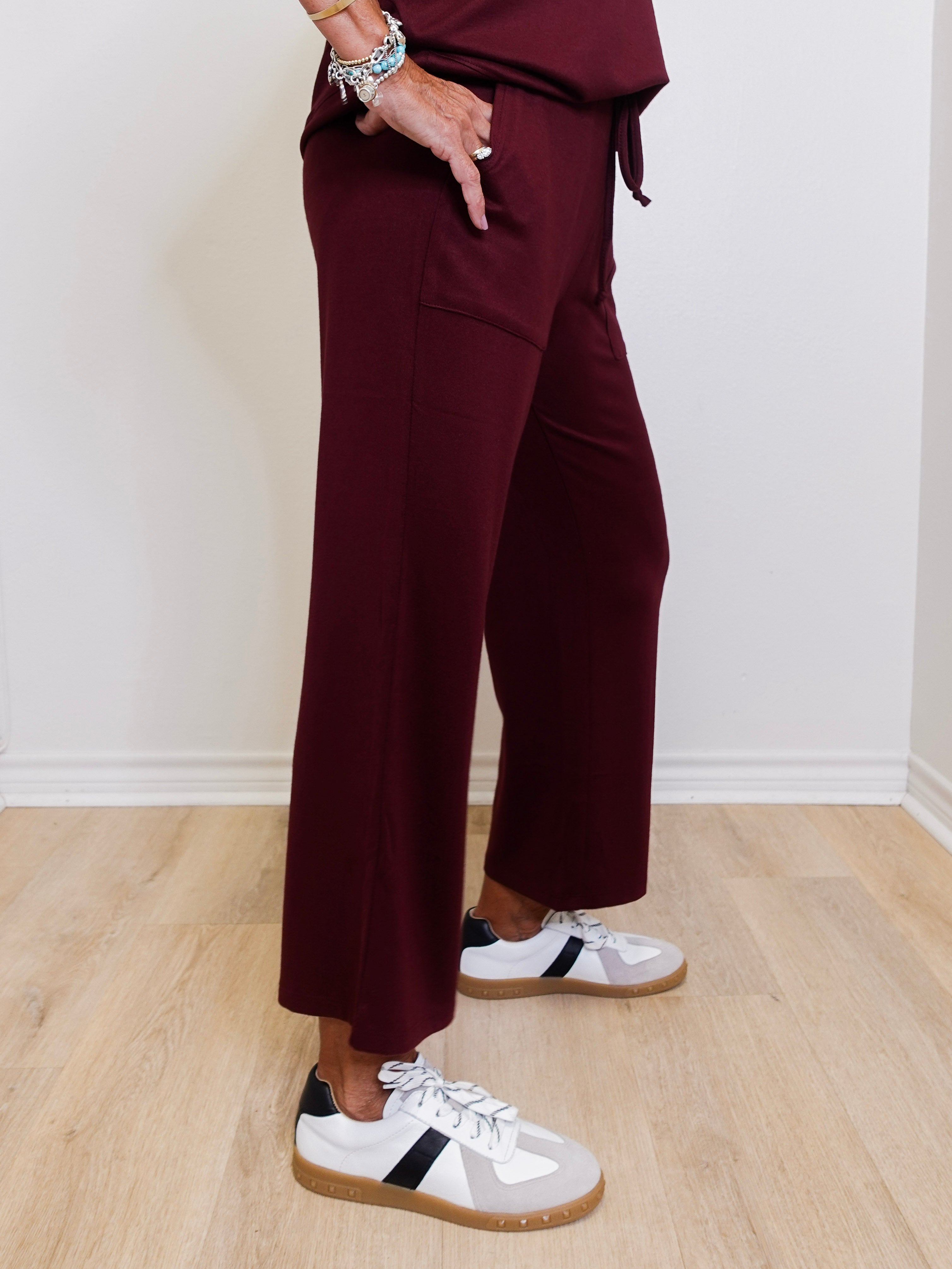 Person wearing burgundy pants and white sneakers with black stripes on a wooden floor.