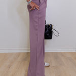 Person wearing a matching purple outfit with wide-leg pants and a black bag on a wooden floor.