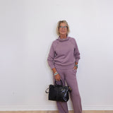 Person wearing a purple outfit holding a black handbag against a white wall.