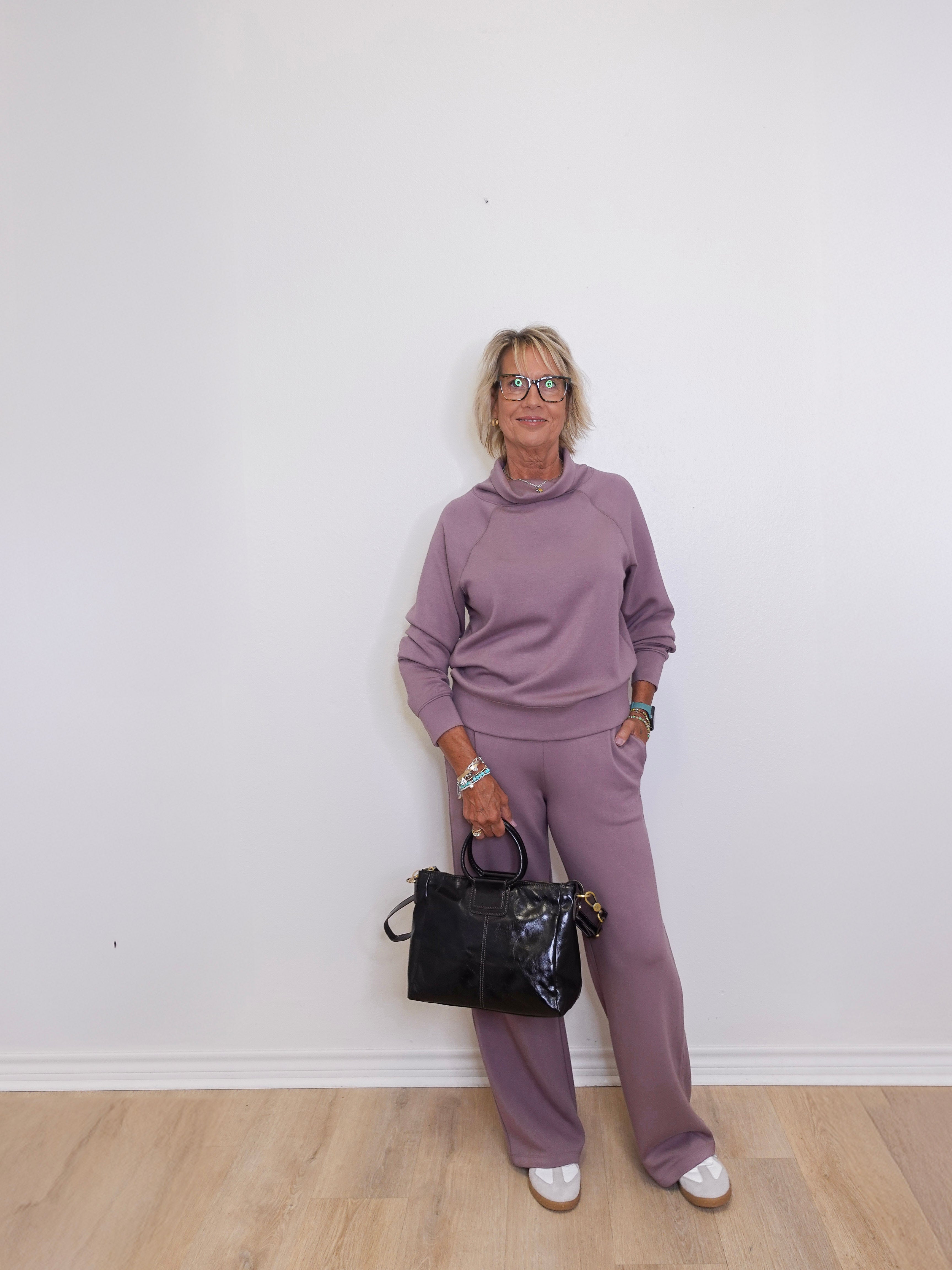 Person wearing a purple outfit holding a black handbag against a white wall.