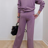 Person wearing a matching purple outfit holding a black handbag against a white wall.