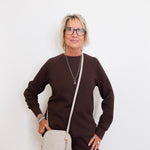 Woman wearing a brown sweater and black pants, holding a beige handbag against a white background