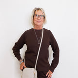 Woman wearing a brown sweater and black pants, holding a beige handbag against a white background