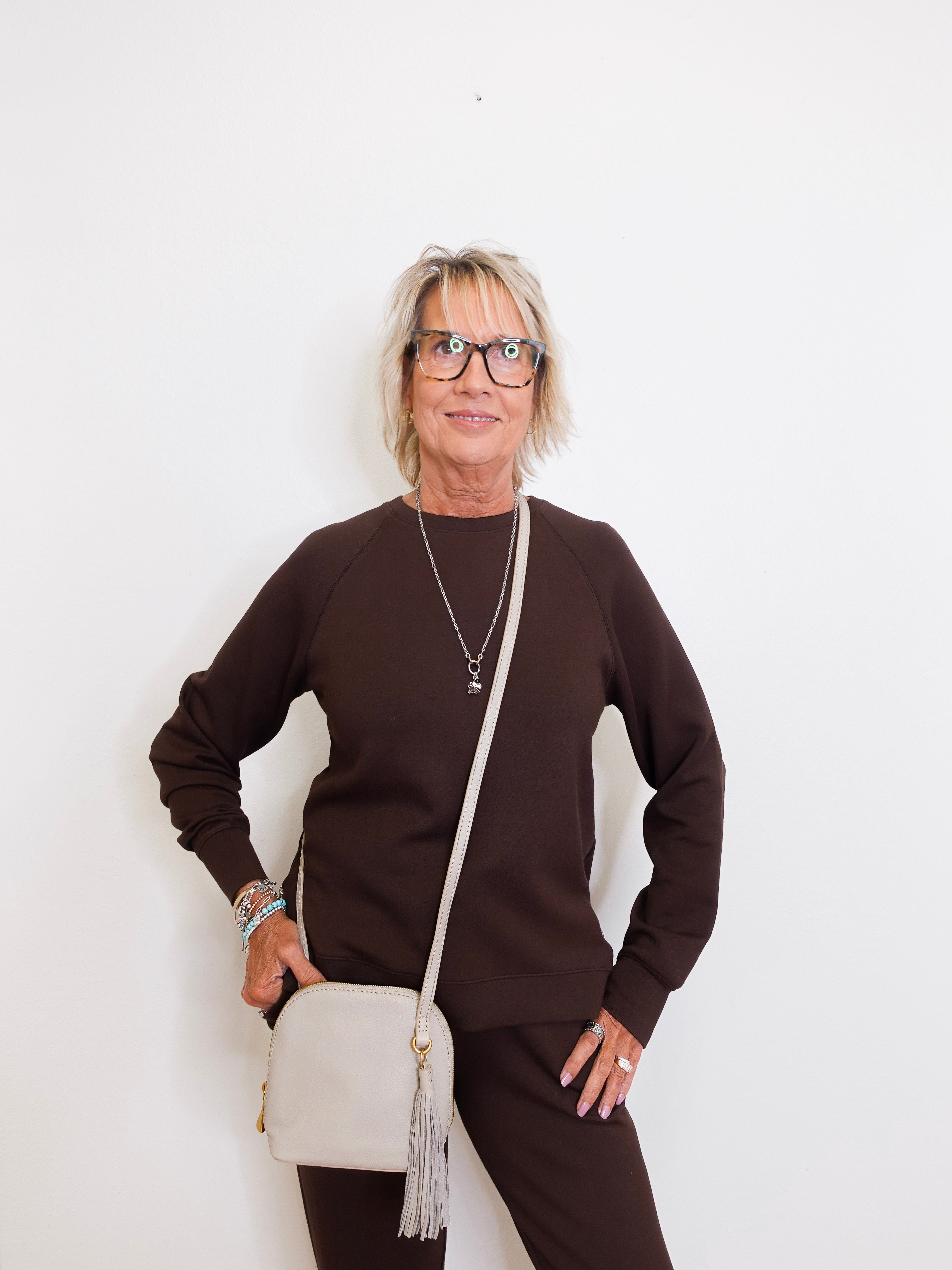 Woman wearing a brown sweater and black pants, holding a beige handbag against a white background