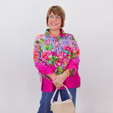 Woman wearing a colorful floral jacket and blue jeans, holding a beige handbag against a white background.