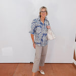 Woman wearing a blue patterned shirt and gray pants against a white wall.