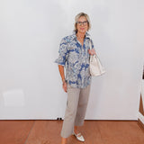Woman wearing a blue patterned shirt and gray pants against a white wall.