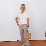 Woman in a white shirt and gray pants holding a beige handbag against a white wall.