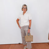 Woman in a white shirt and gray pants holding a beige handbag against a white wall.