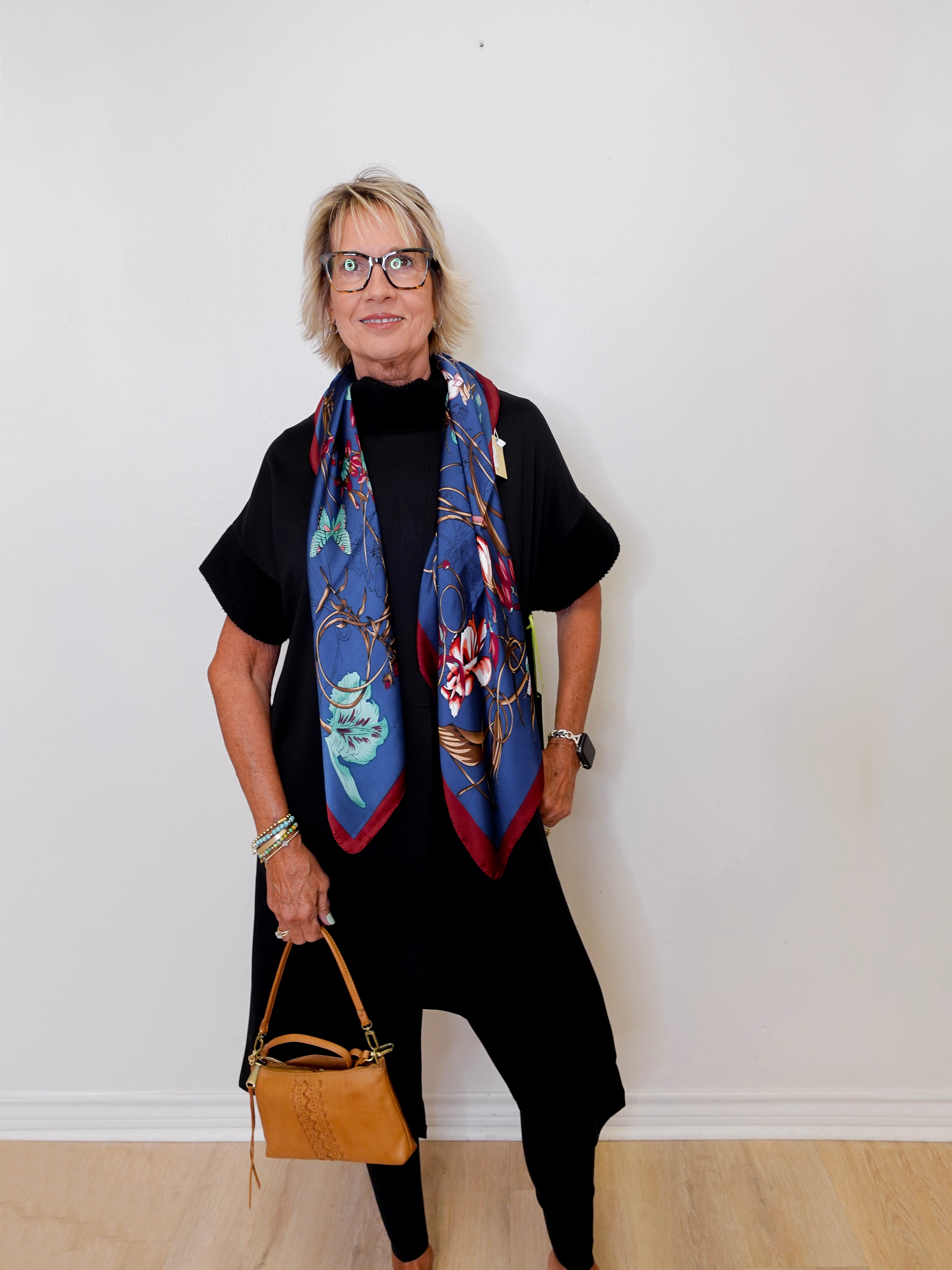 Woman wearing a colorful scarf and holding a brown handbag against a white wall.