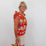 Woman wearing a red floral blouse and beige shorts, holding a beige handbag against a white background.