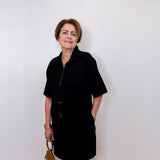 Woman wearing a black dress holding a brown handbag against a white wall.