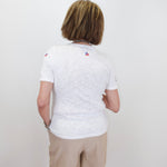Person wearing a white t-shirt with small logos on the back, standing against a plain white wall.