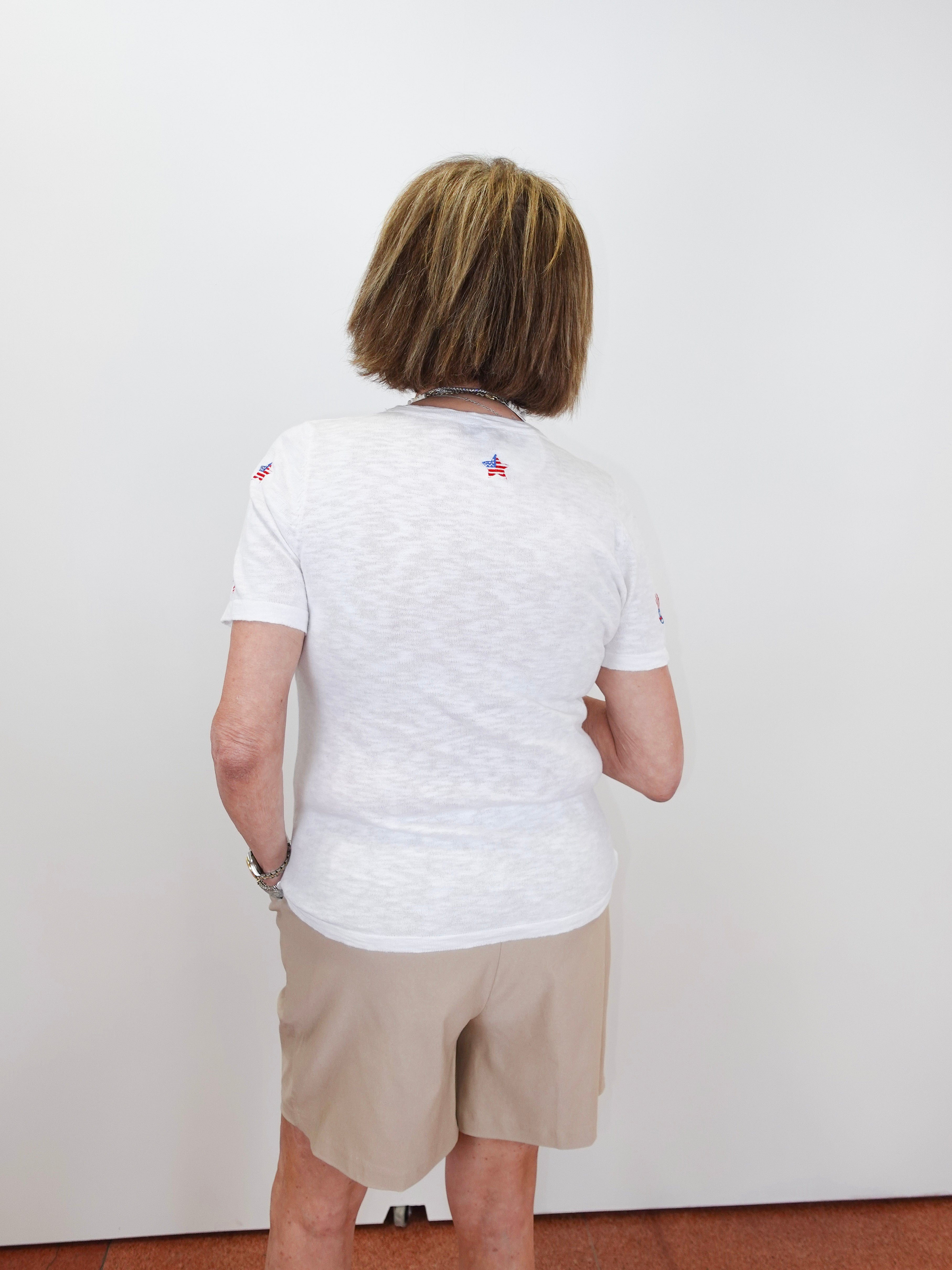 Person wearing a white t-shirt with small logos on the back, standing against a plain white wall.