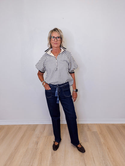 Person wearing a striped shirt and dark jeans standing against a white wall.