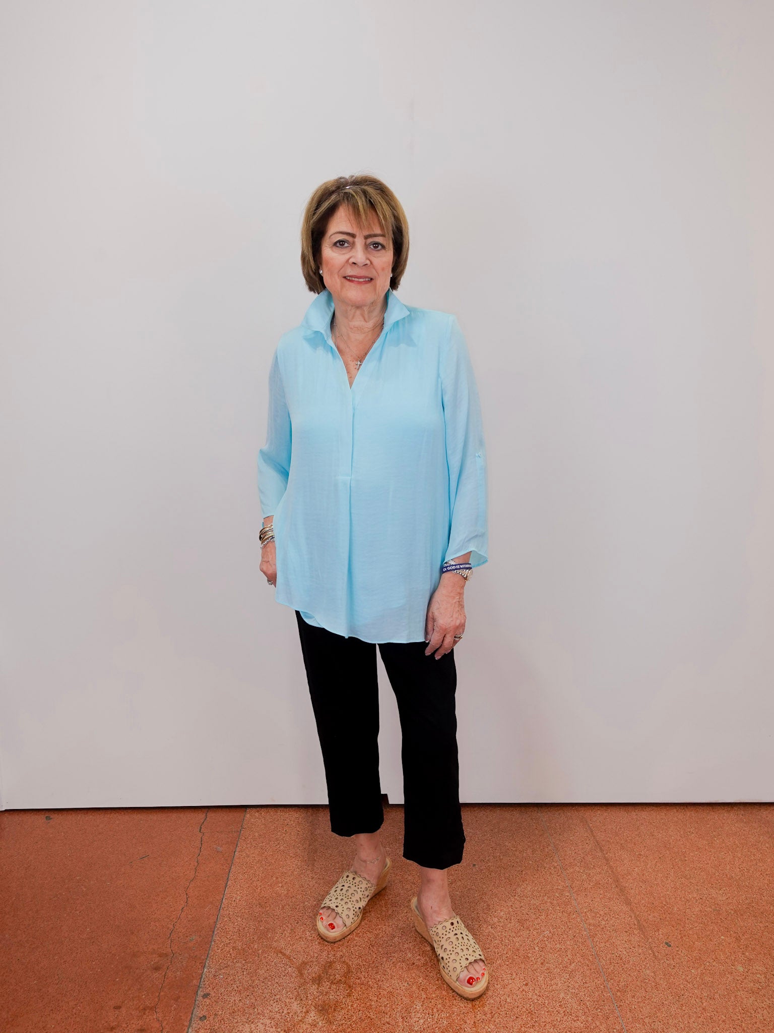 Woman wearing a light blue shirt and black pants standing against a white wall.