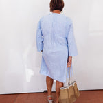 Person wearing a light blue outfit holding a woven bag against a white wall.