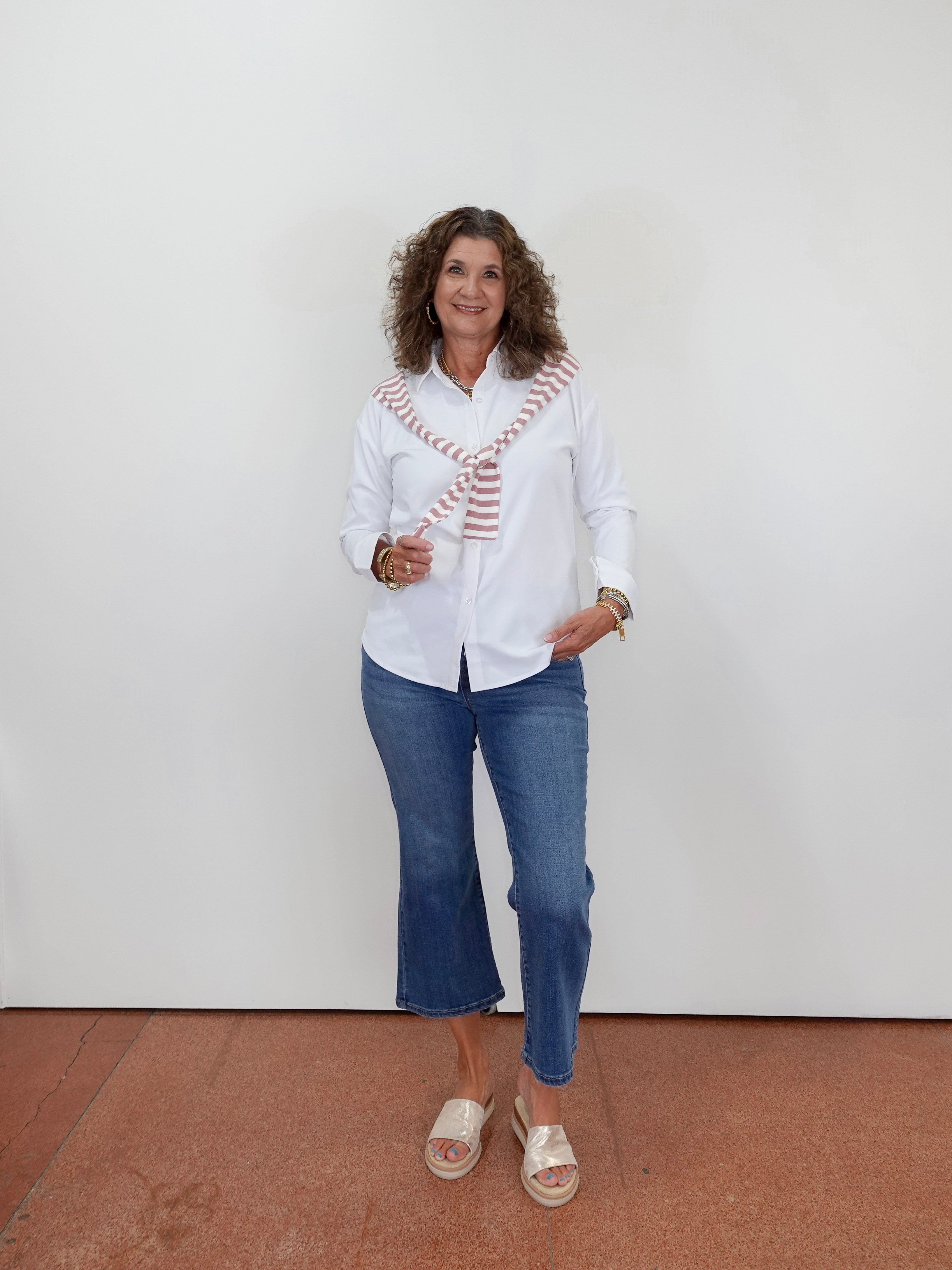 Woman wearing a white blouse with a patterned scarf, blue jeans, and sandals against a white wall.