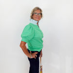 Woman wearing a green blouse with puffed sleeves against a white wall.