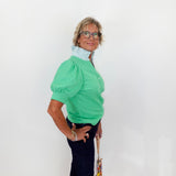 Woman wearing a green blouse with puffed sleeves against a white wall.