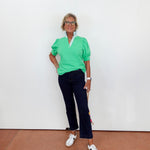 Person wearing a green top and dark pants standing against a white wall.