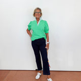 Person wearing a green top and dark pants standing against a white wall.