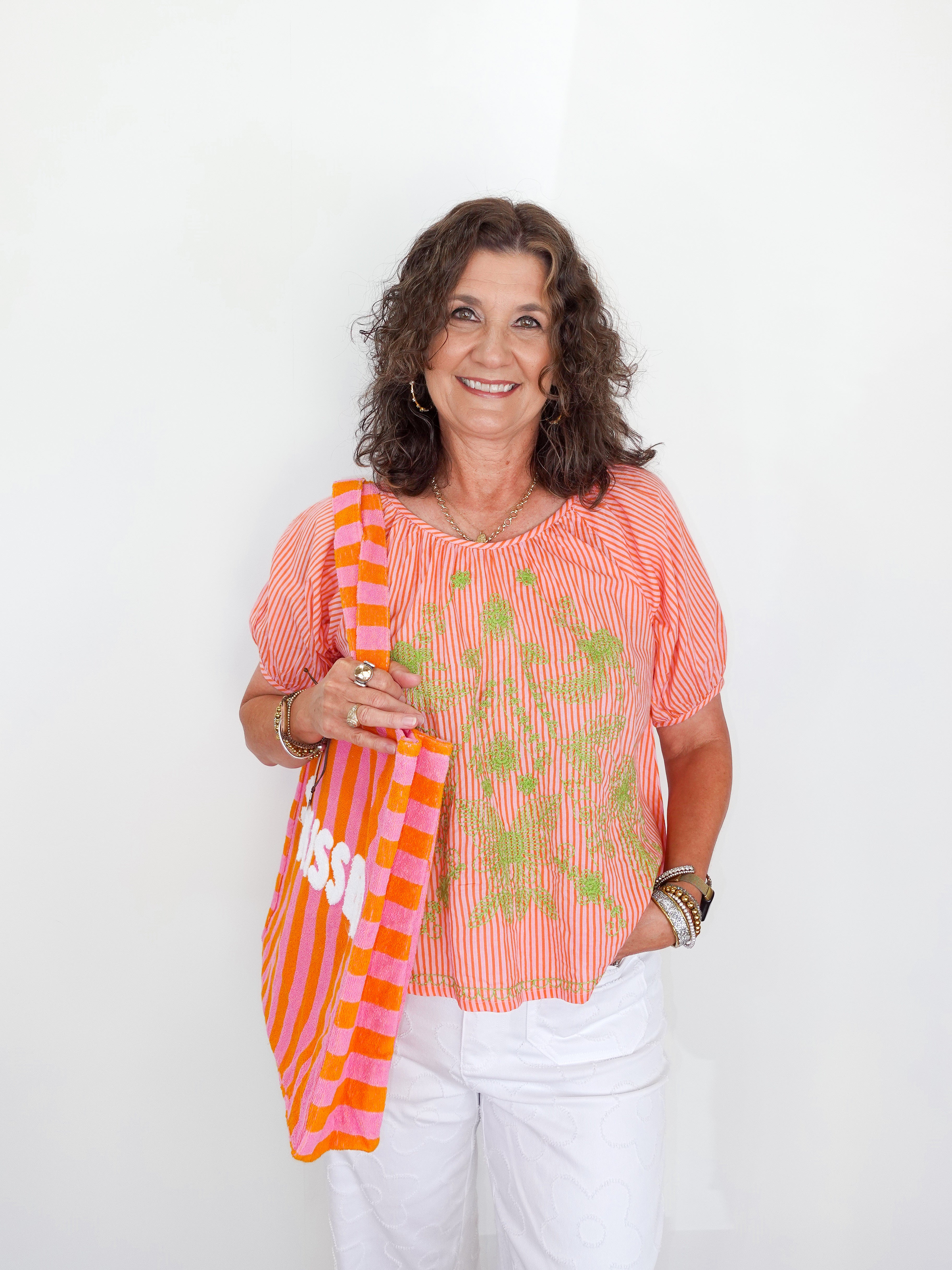 Woman wearing a coral blouse with floral patterns and holding an orange and pink striped bag against a white background